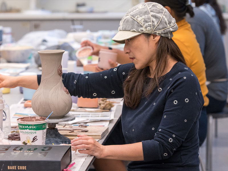 A woman working on a clay vase seated at a table with other class participants