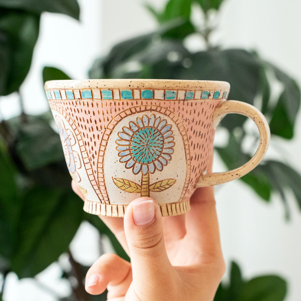 A hand holding a pink ceramic teacup with a blue and green flower