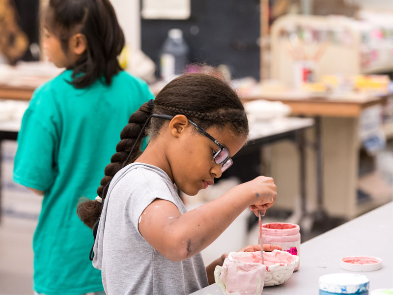 A child with a black braid and glasses glazing pottery