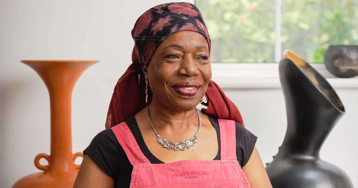 Magdalene Odundo in her studio standing between two vessels and wearing a red apron and headscarf