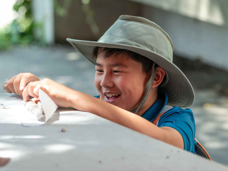 A child working with clay outdoors on a table. He's wearing a large brimmed hat and smiling.