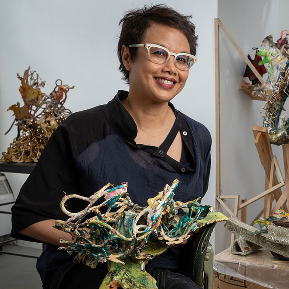 Artist Linda Sormin holding a ceramic sculpture. She is wearing think white glasses and an apron.