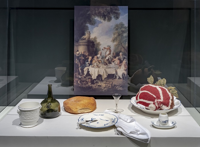 A recreation of an 18th century dinner featuring roast beef, bread, serving plates, and a wine decanter. A painting of a dining scene is in the background